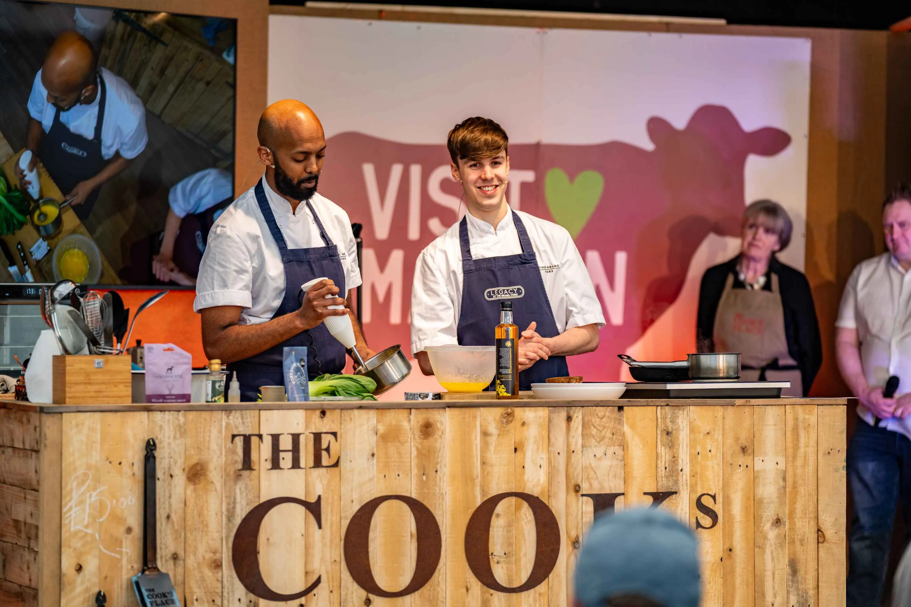 Two chefs presenting a cooking demo at Malton Food festival, with the festival logo behind them